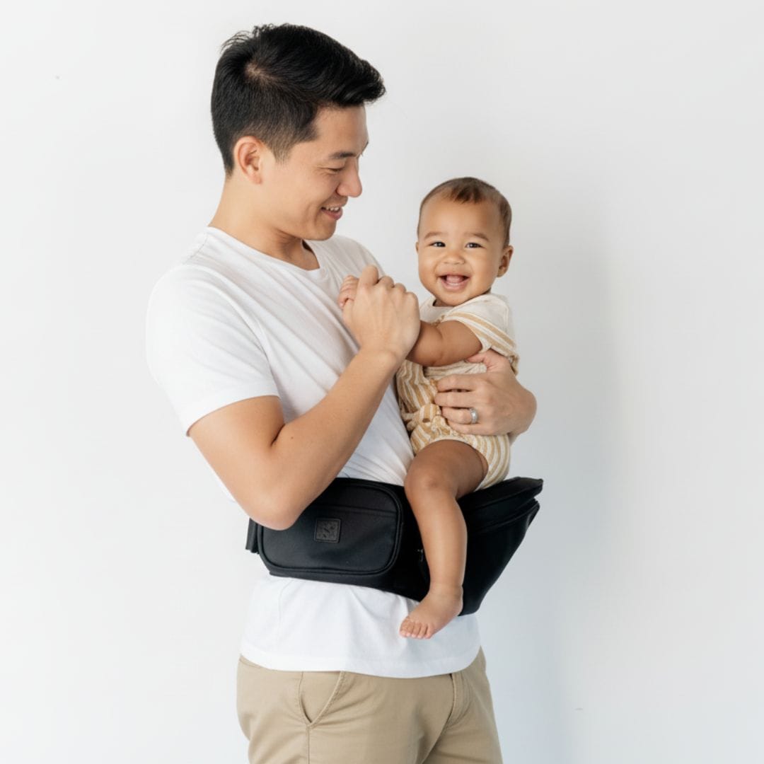 Dad carrying his baby on the classic black HipSurfer hip seat carrier, studio portrait