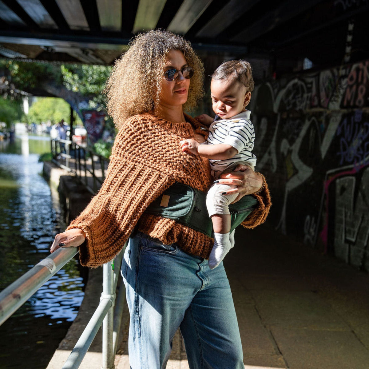 Mum and toddler out for a walk, toddler seated on the moss corduroy HipSurfer hip seat carrier in a graffiti tunnel