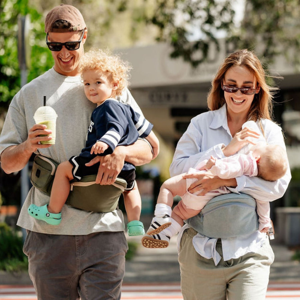 A family of four walking outdoors in a sunny urban setting, each wearing a baby carrier. The image highlights the versatility of the baby carrier with four different babies and toddlers, including a front-facing carrier on the man, a side-mounted carrier on the toddler, a front-facing carrier on the woman, and a side-mounted carrier on the baby.
