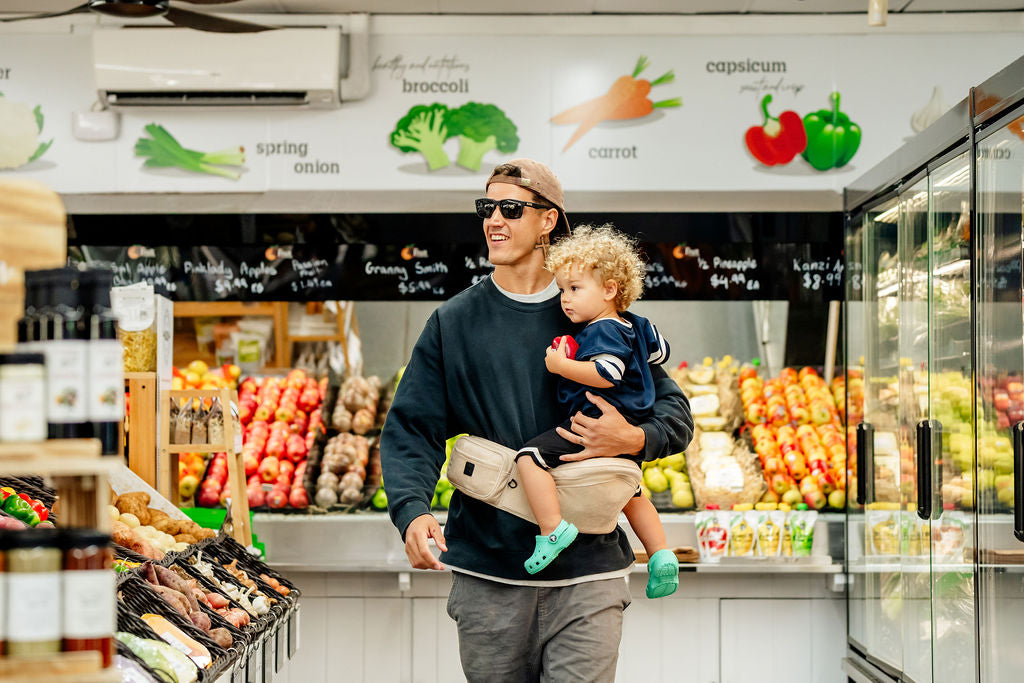 Dad carrying his toddler on a HipSurfer hip seat baby carrier at the fruit shop
