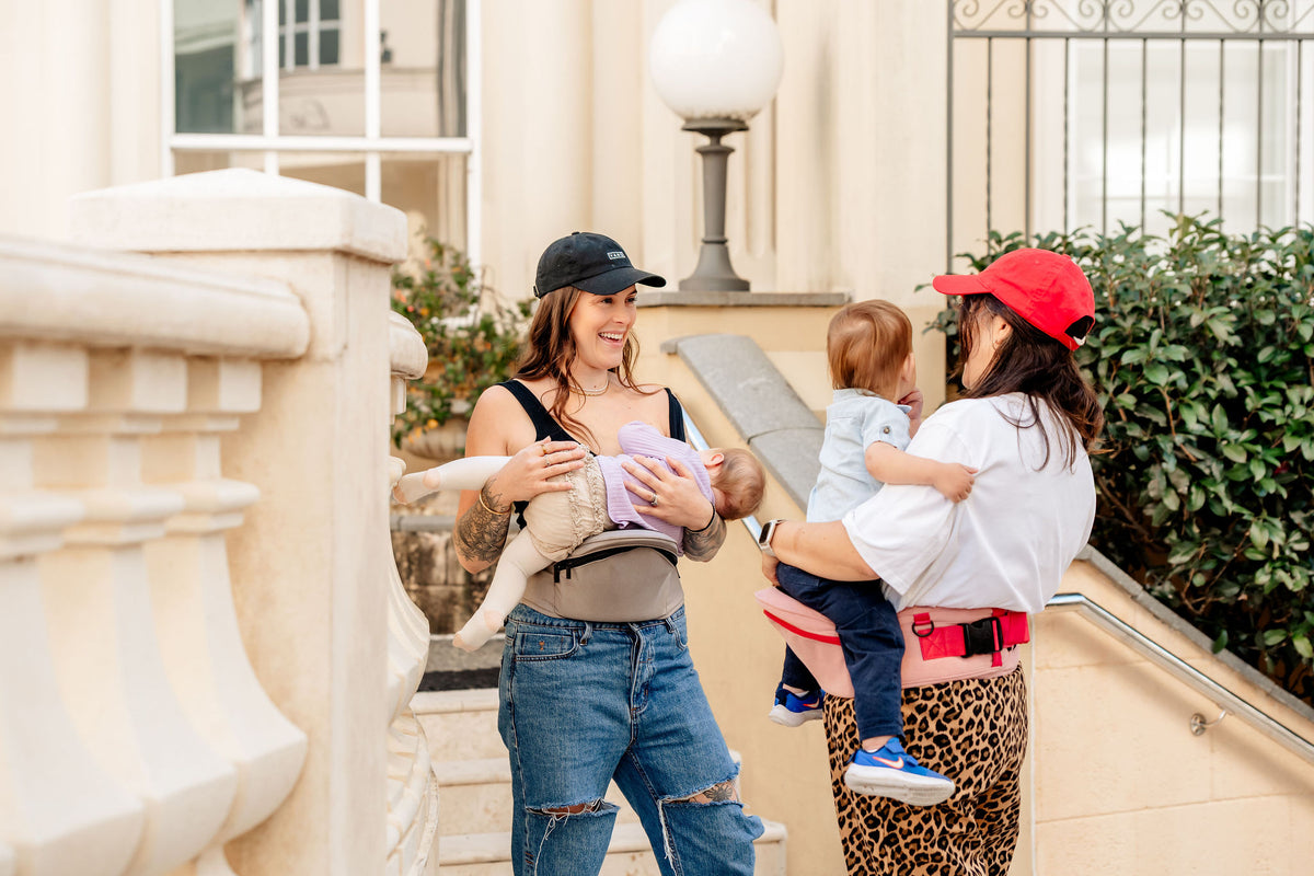 Mum cradling her baby on a grey HipSurfer hip seat baby carrier while chatting to a friend