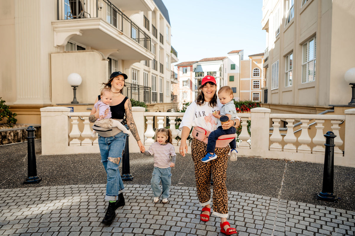 Two mums walking with a toddler, carrying a baby and toddler on grey and pink HipSurfer hip seat baby carriers