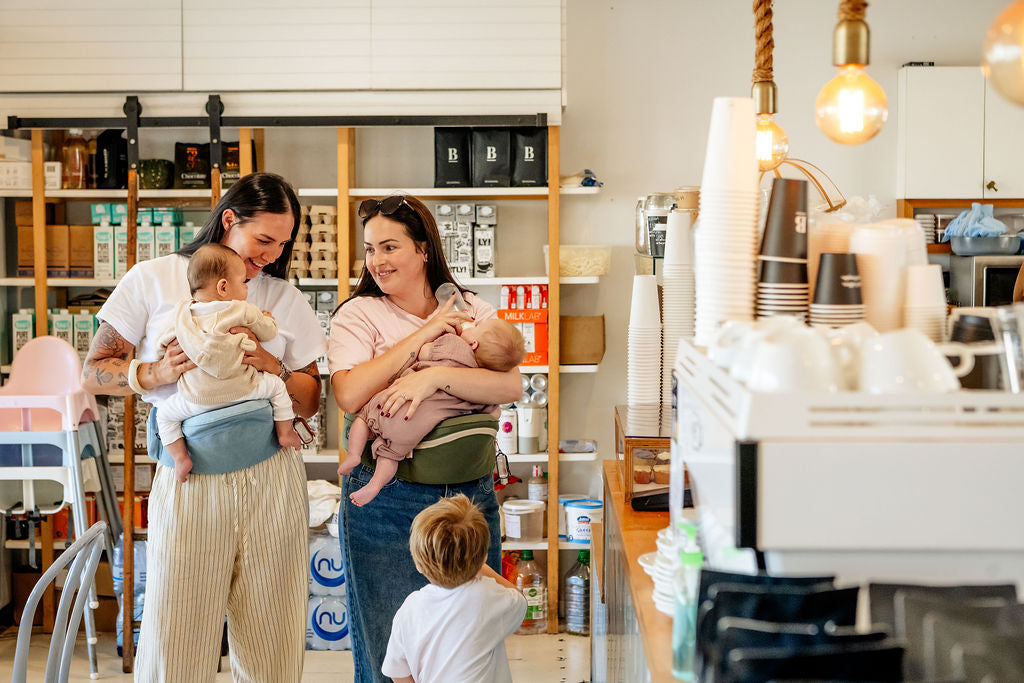 Two mums in a cafe feeding and holding babies on HipSurfer hip seat carriers