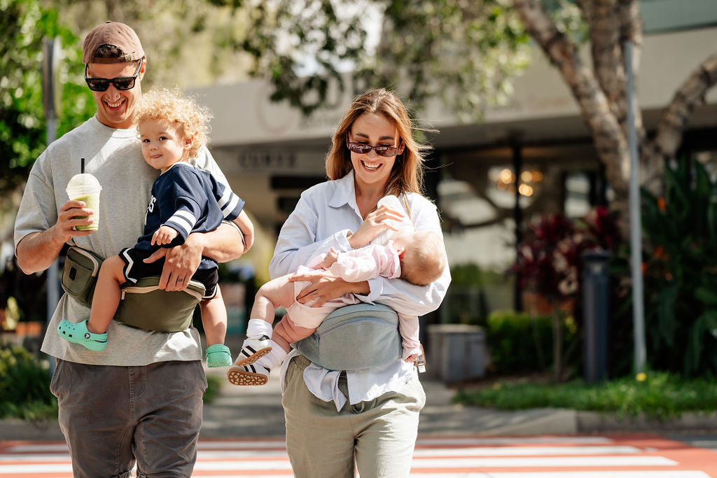 Dad carrying a toddler and mum feeding a baby, both using HipSurfer hip seat baby carriers